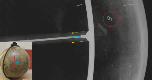 Radiographie du globe terrestre. Dans le cercle rouge, une petite pièce métallique en forme de « a » tout près de (sur ou dans) la calotte sud. Les traits sont de petits clous très fins, invisibles depuis la surface du globe; il est difficile de déterminer si ces clous sont originaux ou non. La ligne bleue indique la position de l'axe métallique et le rectangle rouge la zone du film. Entre les flèches jaunes, se trouve l'axe de bois. © SIK|ISEA